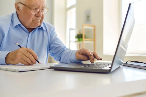 An elderly man wearing a light blue collared shirt sits at a laptop holding a pen and paper is debating between a 401a vs 401k retirement plan.